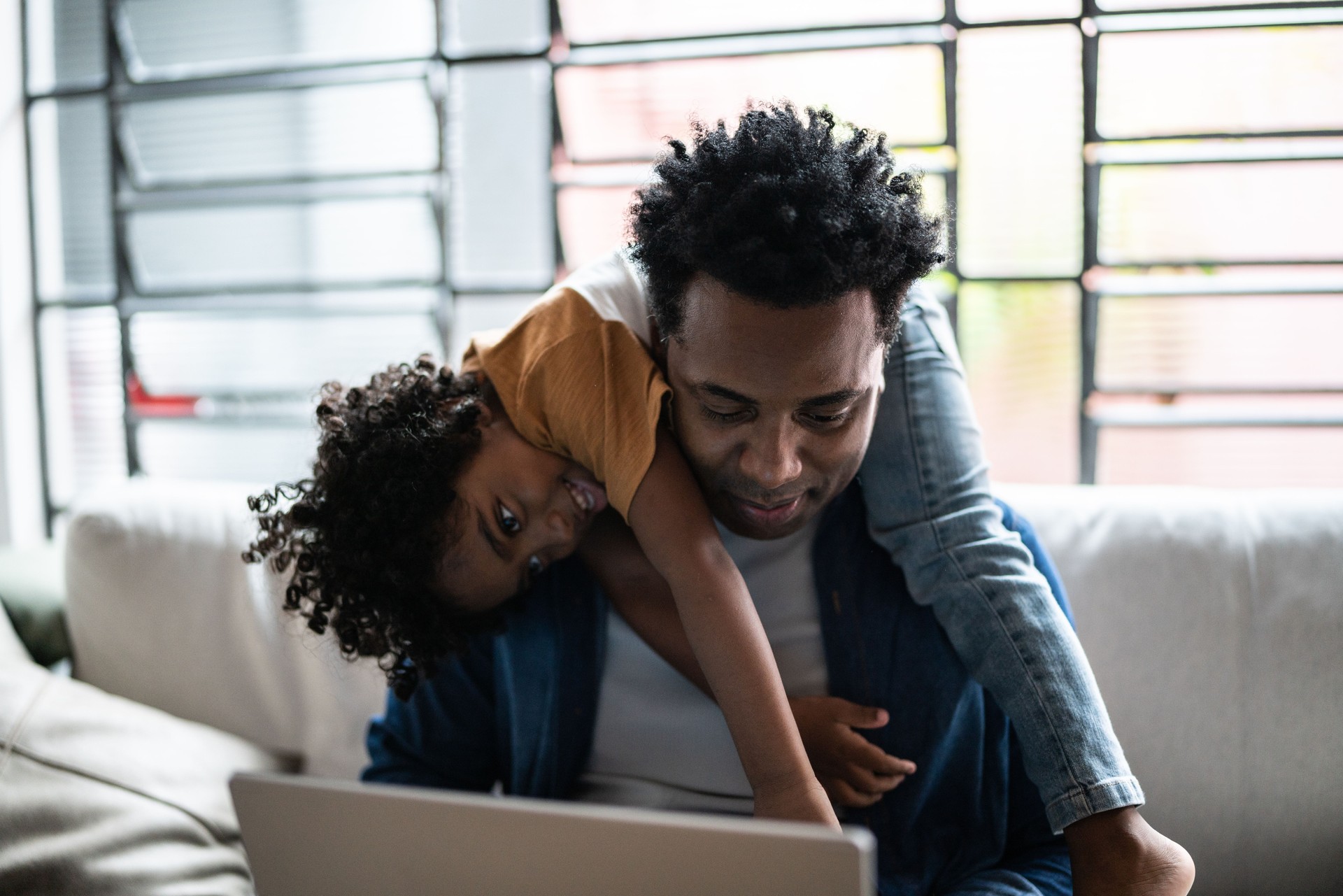 Father using the laptop trying to work while son is on his back at home